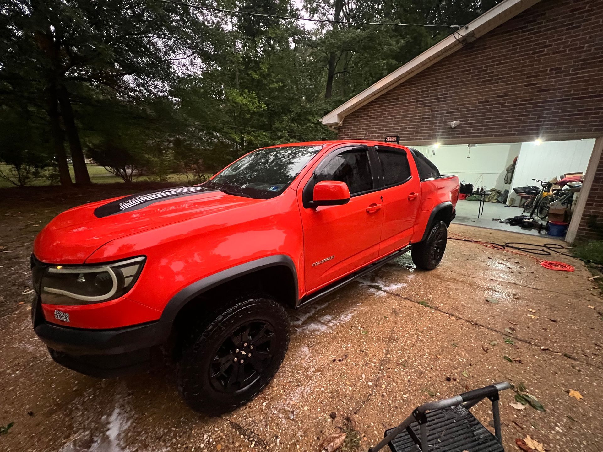 Red pickup truck parked on a wet driveway beside a garage, with another vehicle and trees in the background