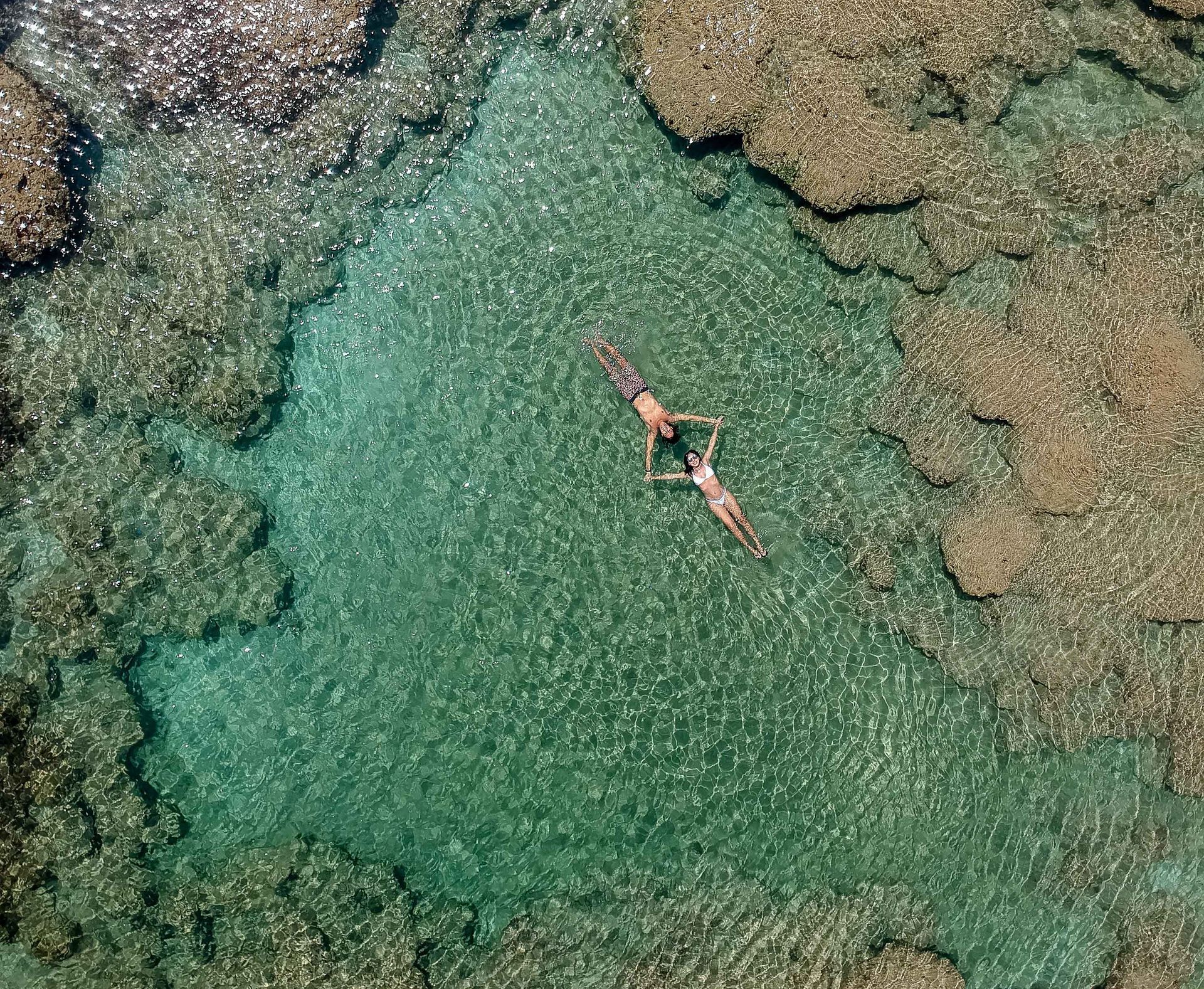 Duas pessoas flutuam de bruços em águas cristalinas azul-turquesa cercadas por formações rochosas.