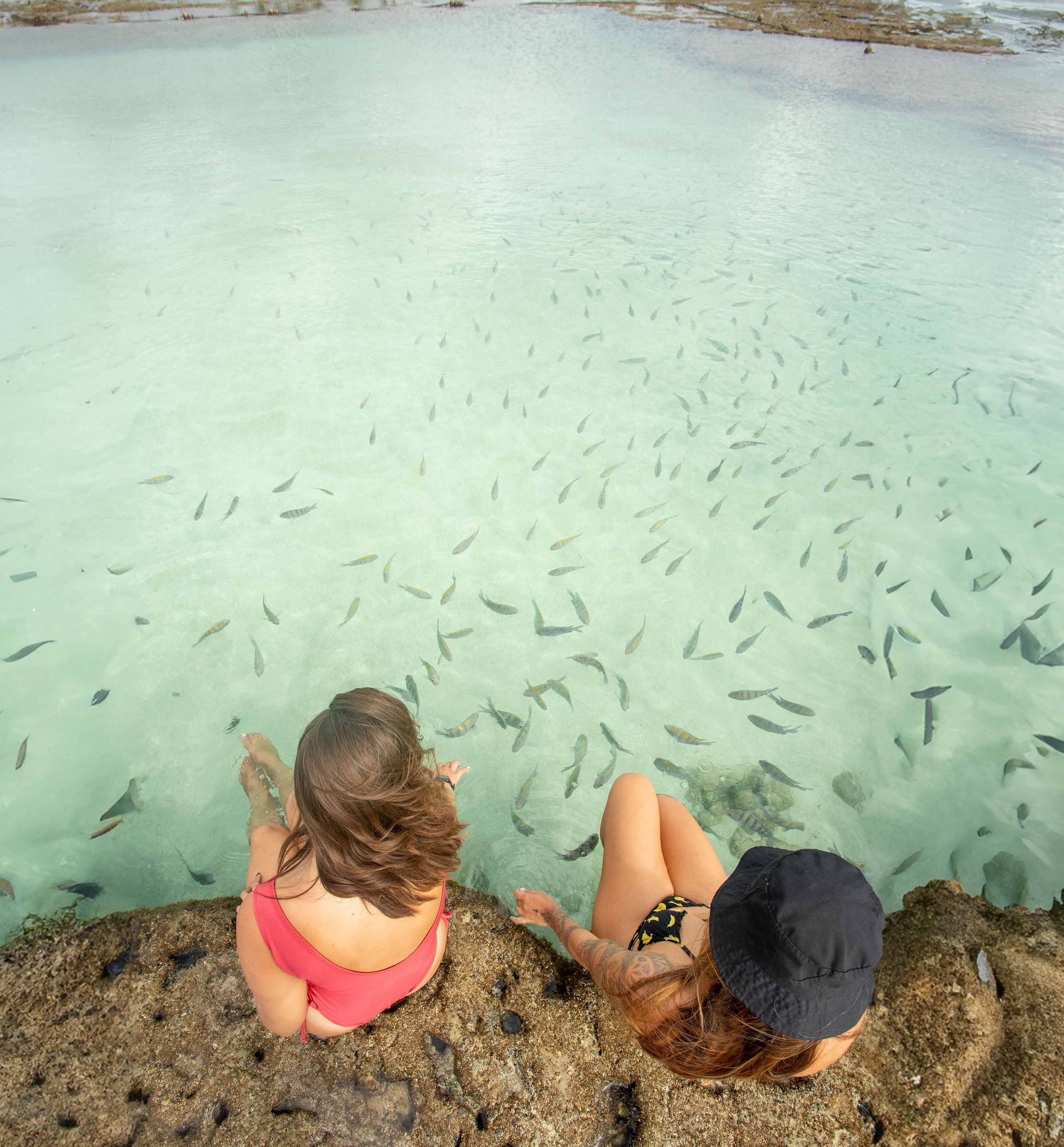 Casal relaxando na praia, oceano com veleiros ao fundo.