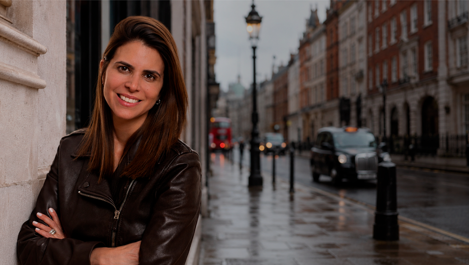 Smiling woman in a brown leather jacket leaning against a wall on a wet city street.