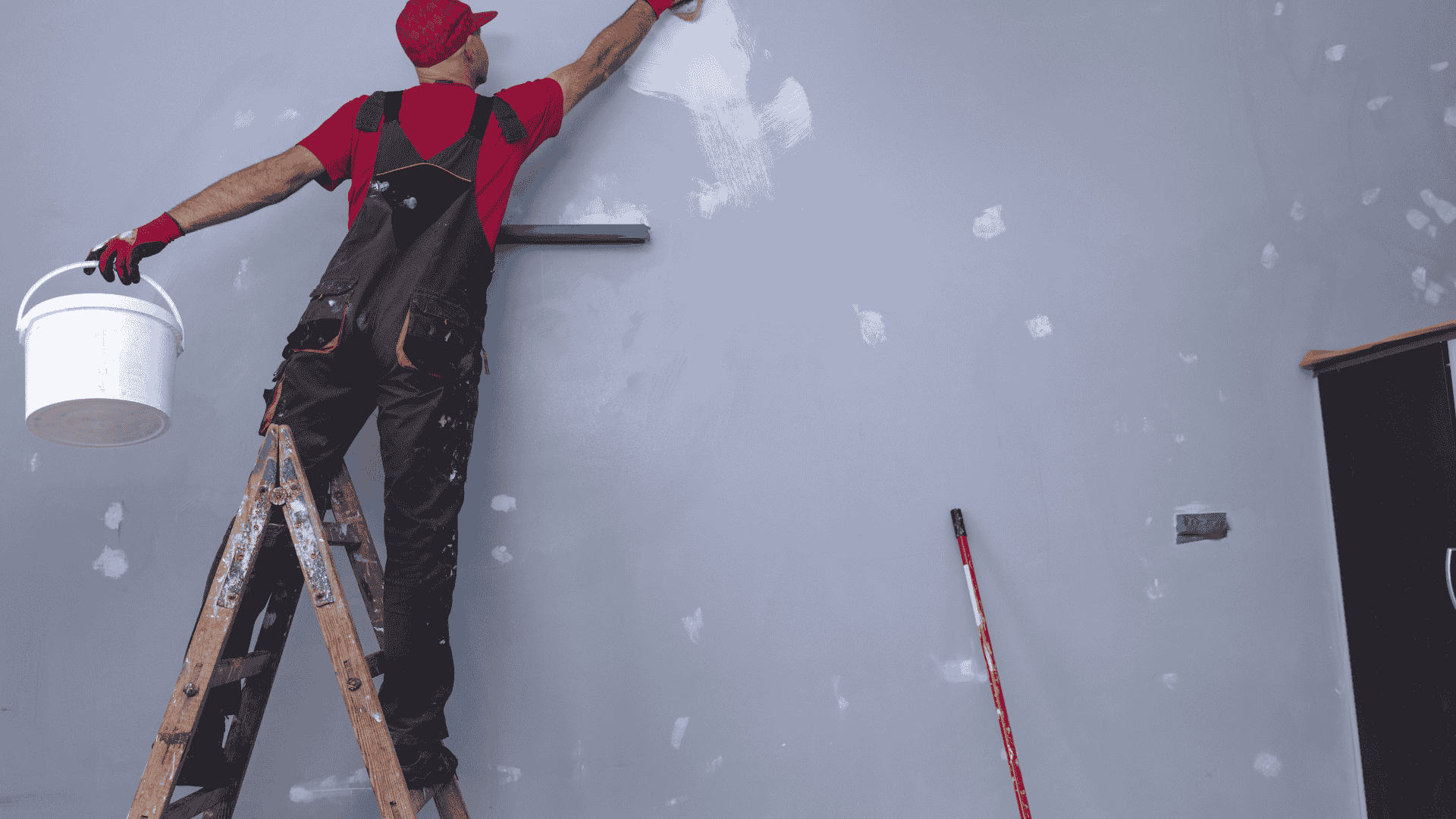 Painter on a ladder applying white paint to a wall with a roller and bucket in a room under renovation
