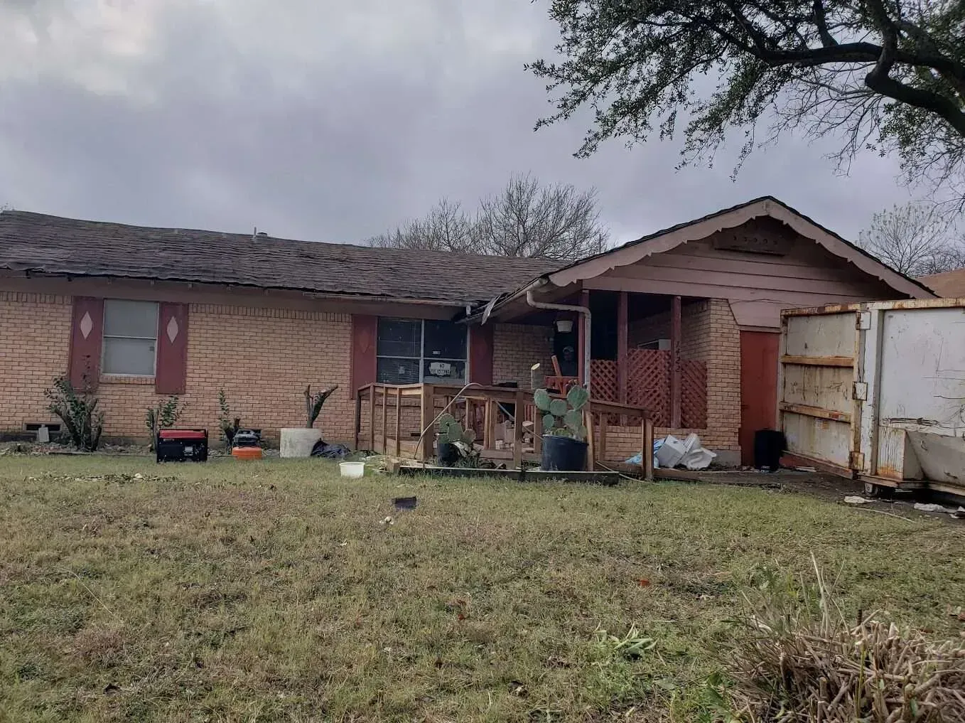 Single-story brick house with damaged roof, boarded window, and cluttered front yard under cloudy sky