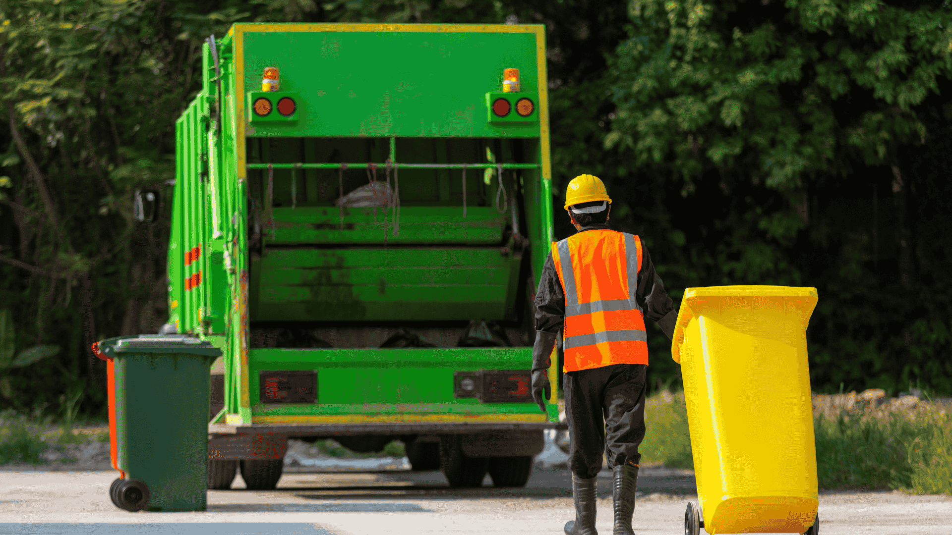 Worker in orange vest stands behind a green garbage truck with yellow bins on a road