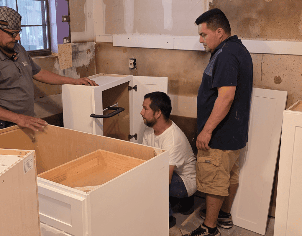 Three people assembling white kitchen cabinets in a room under renovation.