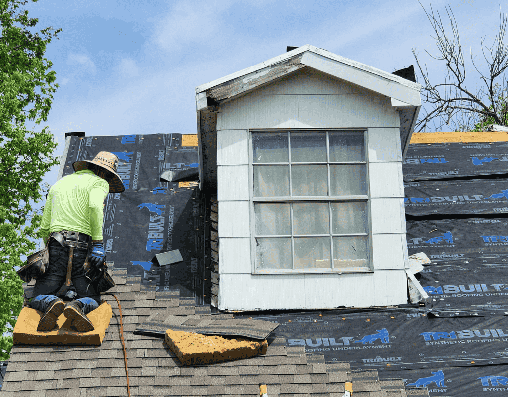 Worker roofing a house dormer, installing black underlayment around a white window on a sloped roof.