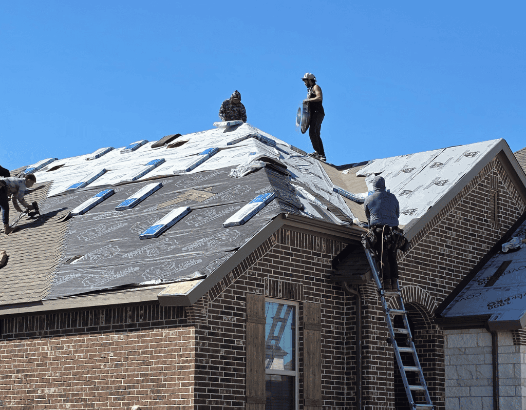 Workers on a brick house roof removing snow under a clear blue sky.