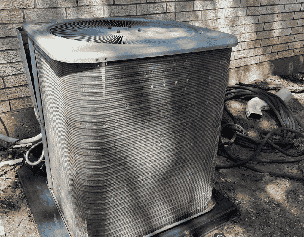 Outdoor air conditioner unit with a metal grille, sitting on a concrete pad near coiled hoses and a wall.