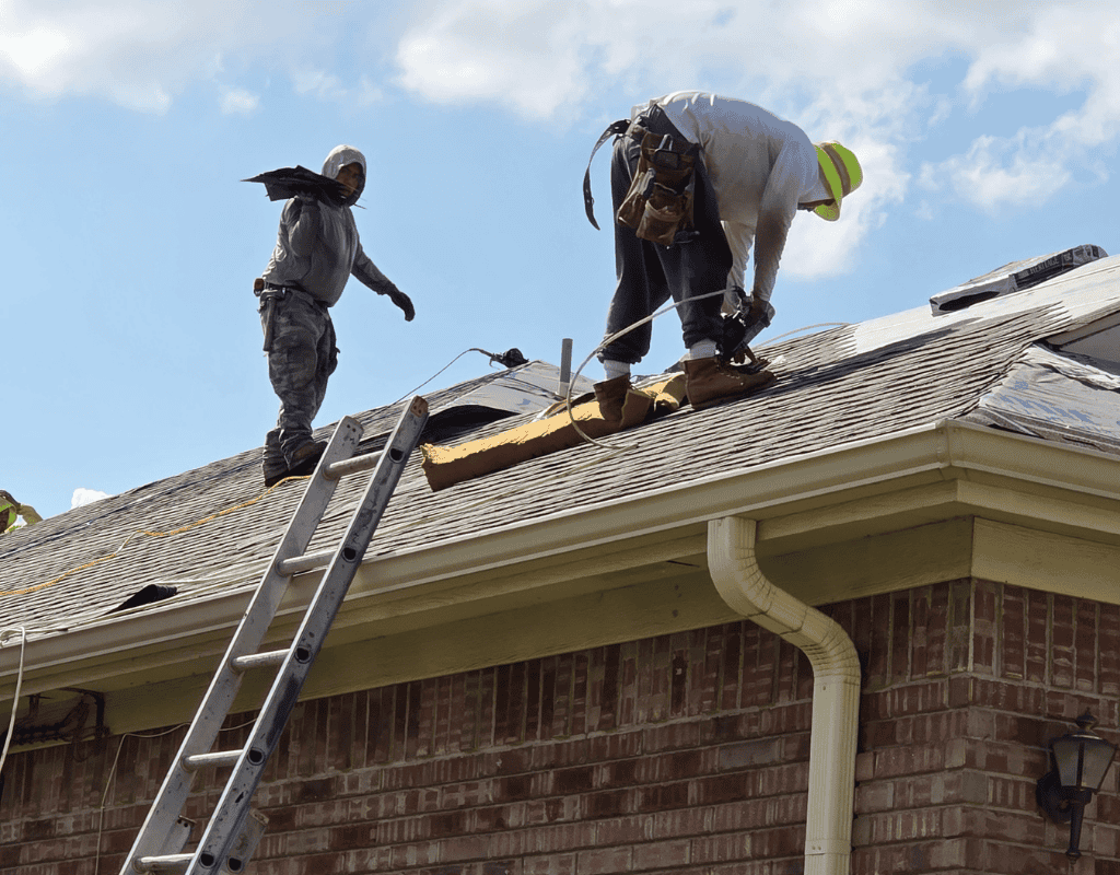 Two workers repairing a roof, one on a ladder and one kneeling near tools under a blue sky