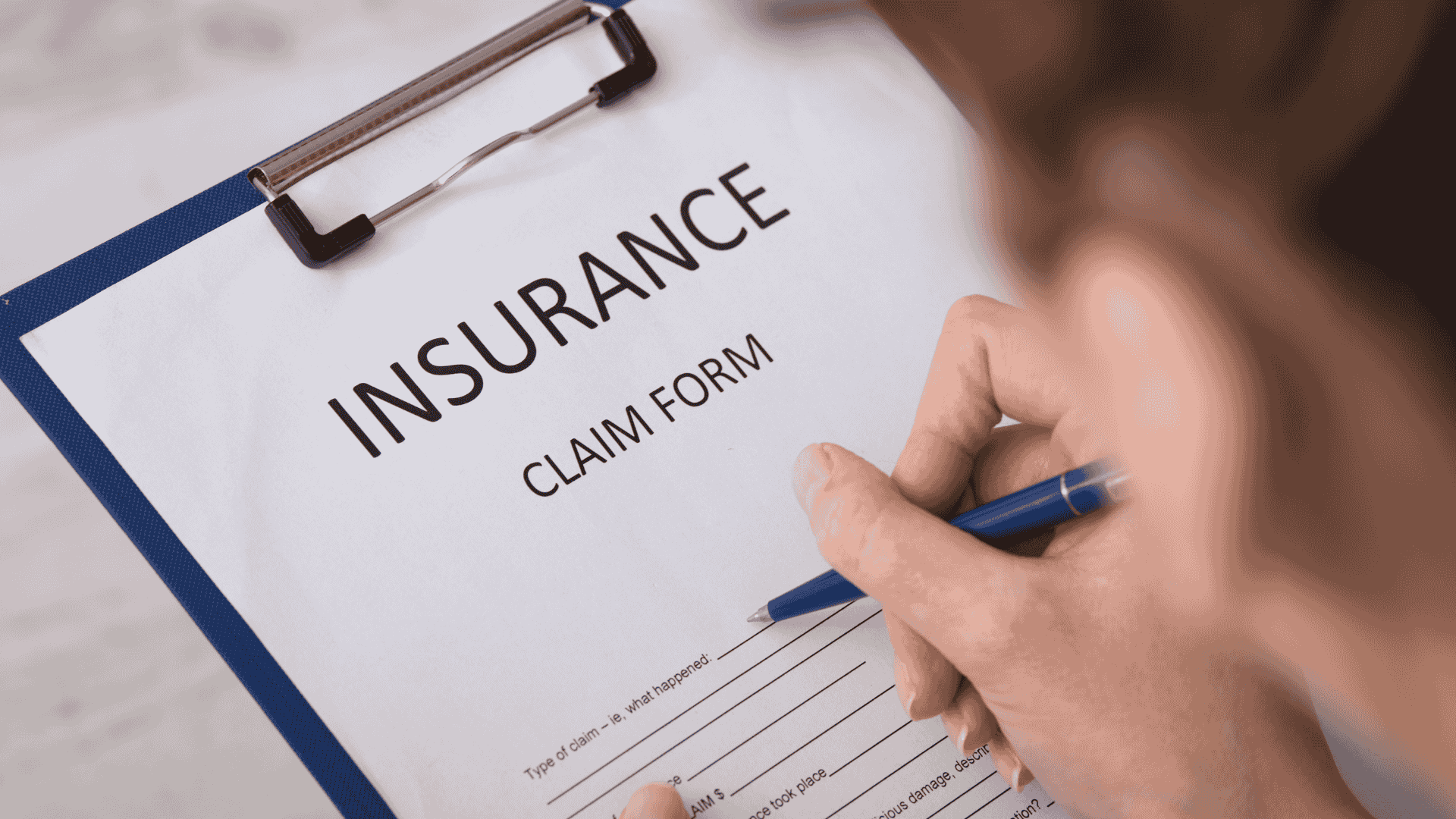 Hand filling out an insurance claim form on a clipboard with a blue pen