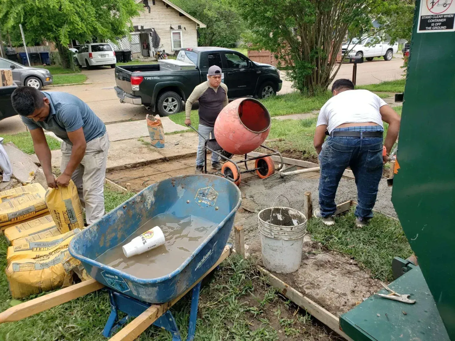 Workers mixing concrete on a driveway with a wheelbarrow, cement mixer, and buckets
