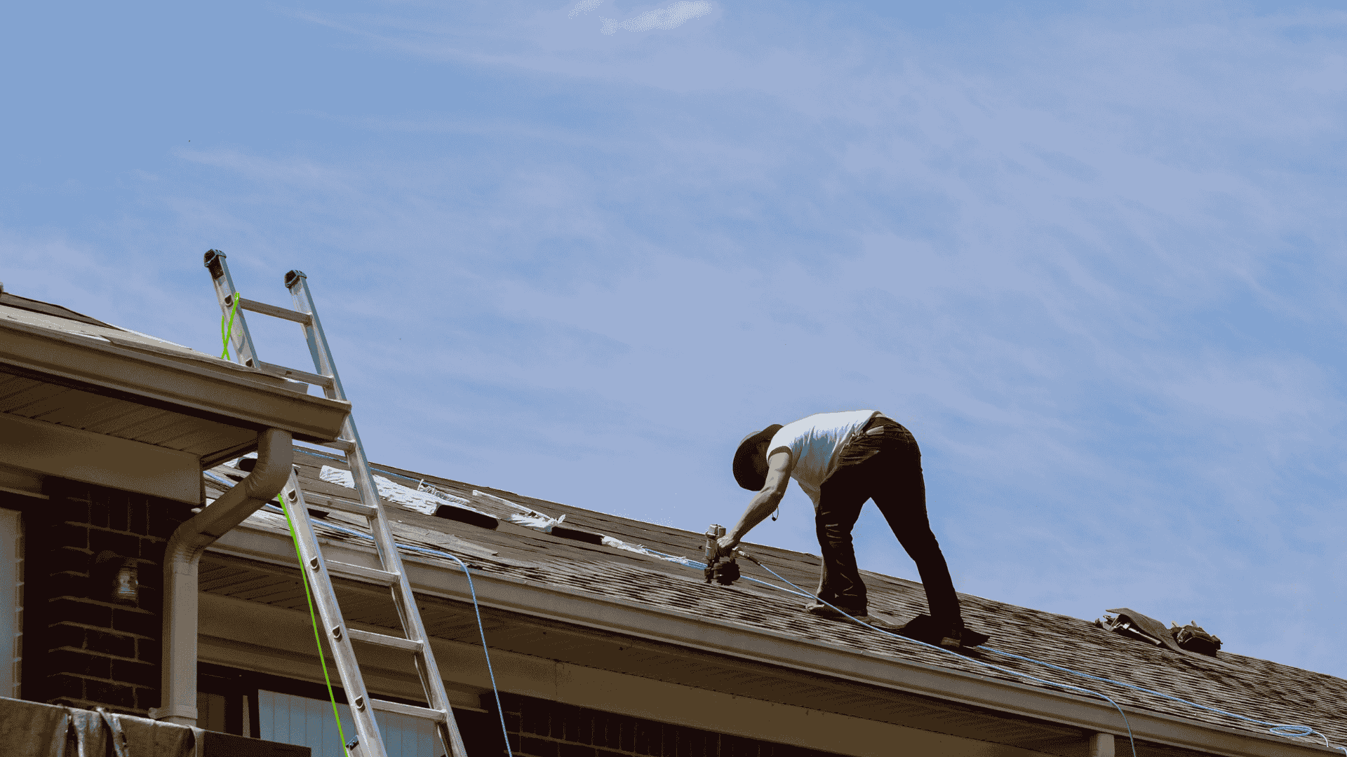 Worker on a roof with a ladder, repairing shingles under a blue sky.
