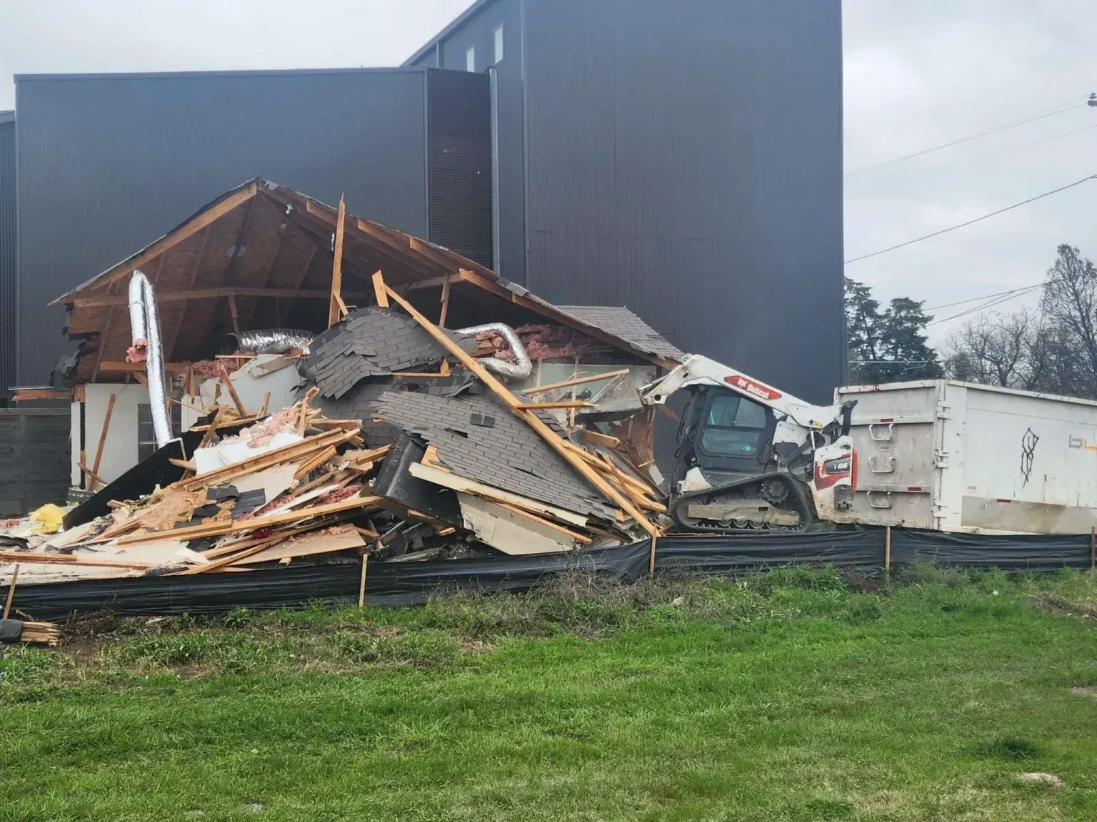 Collapsed building debris beside a dark warehouse, with a white dumpster and grass in front