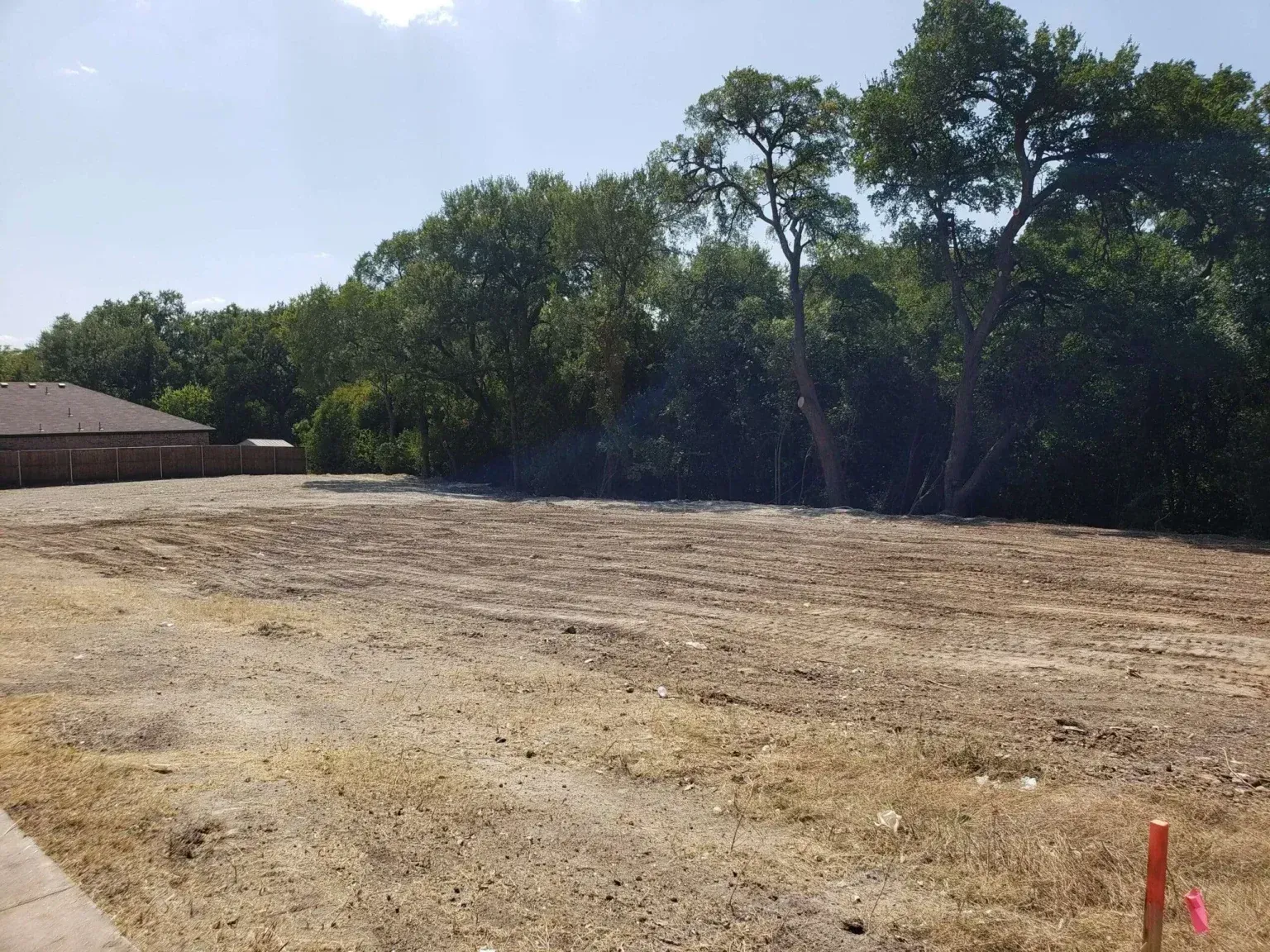 Cleared dirt lot beside trees and a brick building under a bright sky
