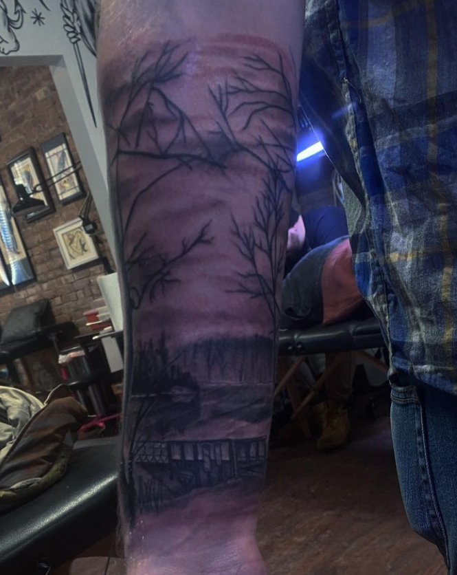 Arm tattoo with a dark forest and cabin silhouette on a forearm, indoors.