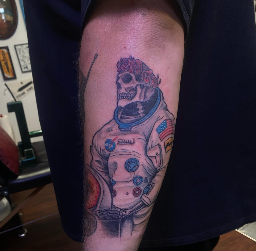 Close-up of a tattooed upper arm showing an astronaut skull design with an American flag patch.