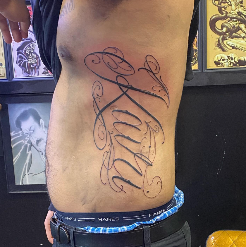 Side torso with large cursive black tattoo, standing indoors against framed wall art.