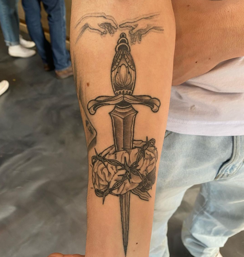 Forearm tattoo of a sword with a floral hilt and wings near the top, in black ink