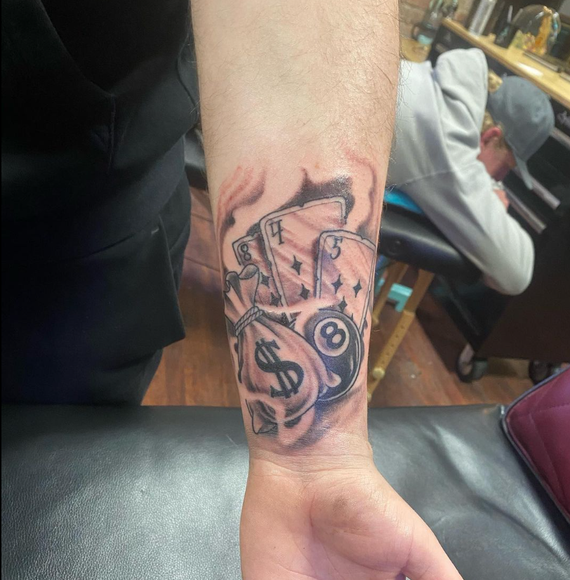 Forearm tattoo of an 8-ball with cash and playing cards in black and gray ink