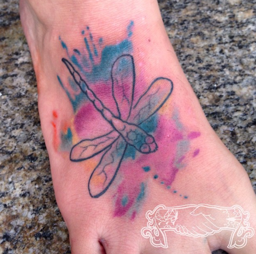 Dragonfly tattoo with pink, blue, and teal watercolor splashes on a foot/ankle.