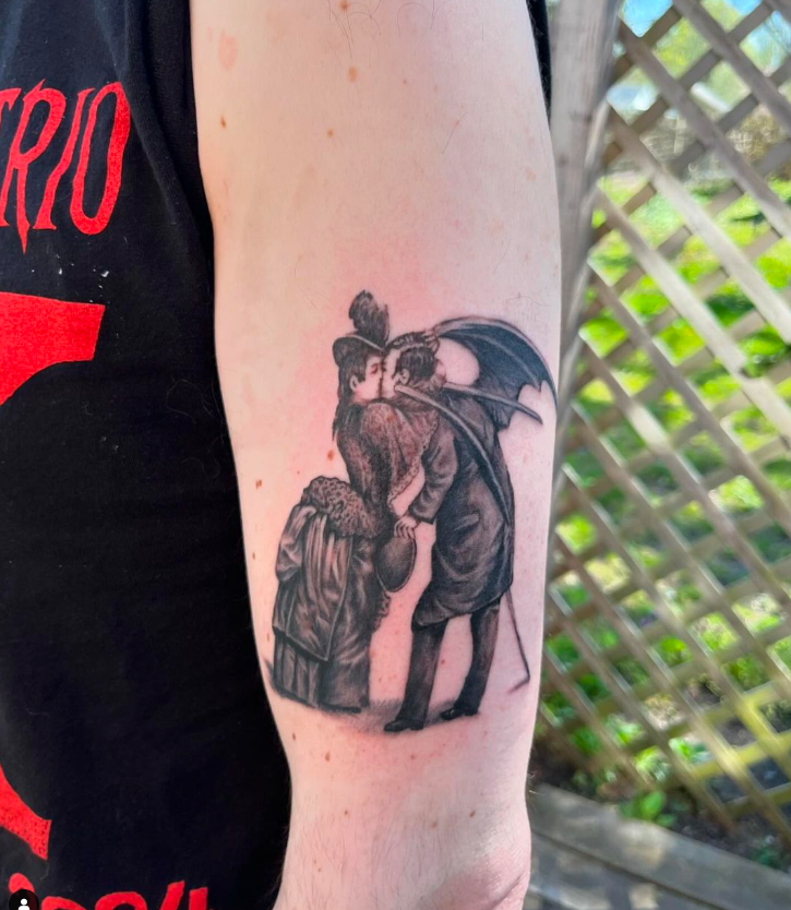 Black tattoo of two figures embracing on a person’s upper arm, with a lattice fence in the background