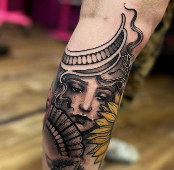 Detailed black-and-gray tattoo of a serene face with a headdress and yellow flower on a leg