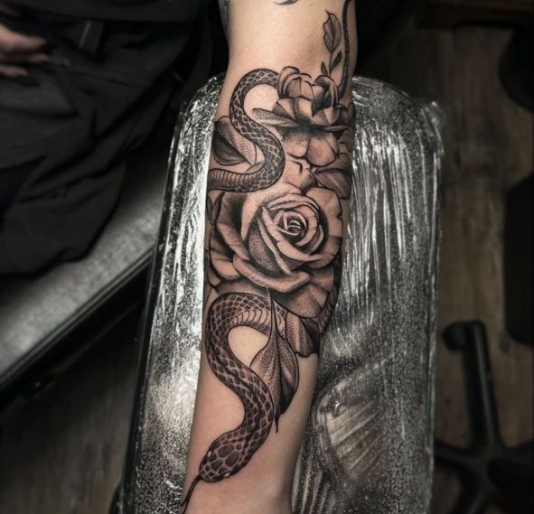 Snake and rose tattoo sleeve on a forearm, black and gray ink, resting on a chair.
