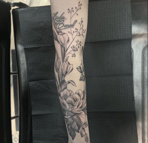 Black-and-gray floral tattoo sleeve on a person’s forearm against a dark seat background