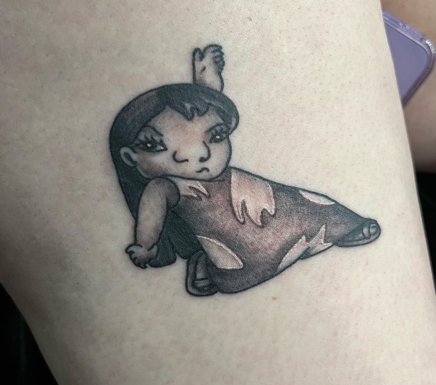 Small black-and-gray cartoon-style tattoo of a seated girl on skin, with a tilted head and skirt-like dress