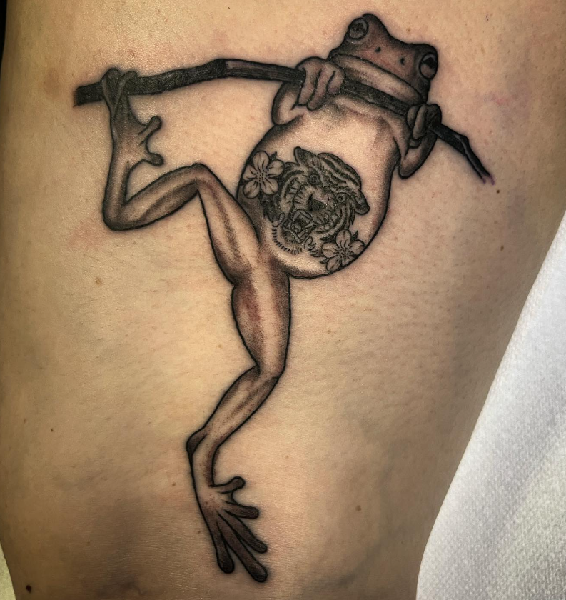 Black-and-gray tattoo of a frog hanging from a branch, with a circular design on its belly