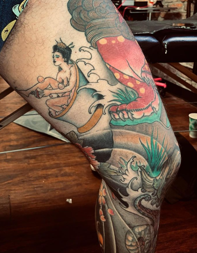 Colorful leg tattoos on a bent leg, featuring a seated figure, koi fish, waves, and floral details.