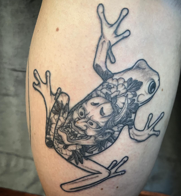 Black-and-gray tattoo of a frog with a detailed floral-and-face design on its body