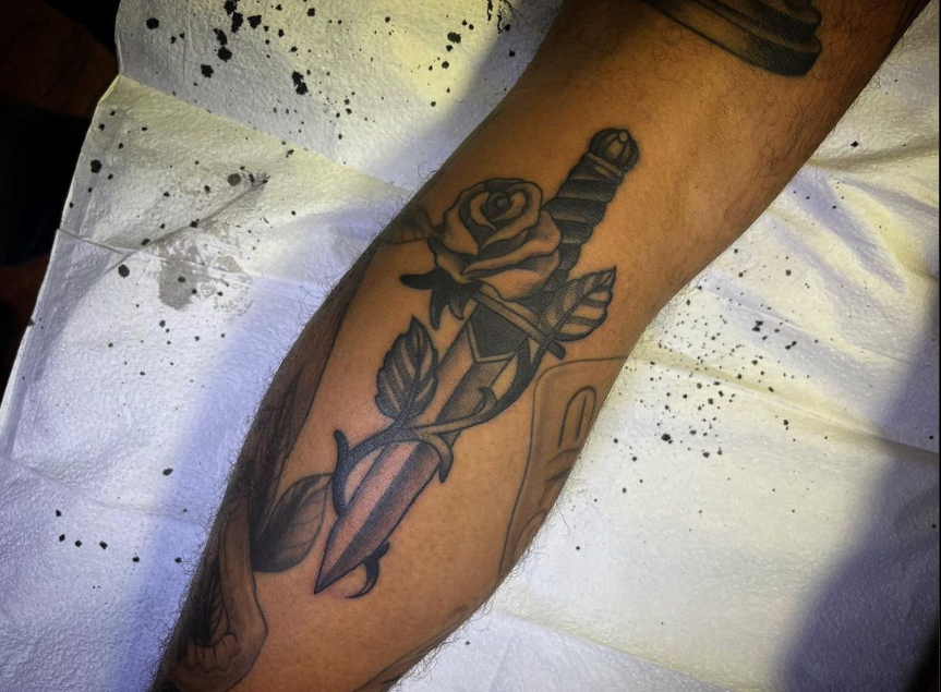 Black rose tattoo on a forearm, with leaves and a long stem against white bedding