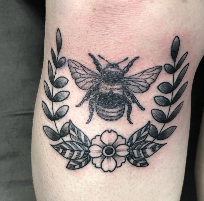 Black bee tattoo surrounded by laurel leaves and a flower on skin