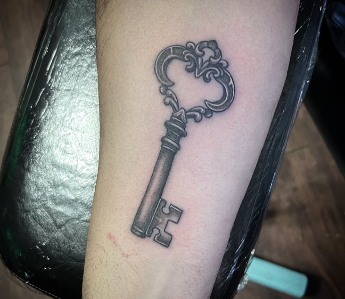 Small black key tattoo on a person’s forearm, with ornate decorative details on the bow.