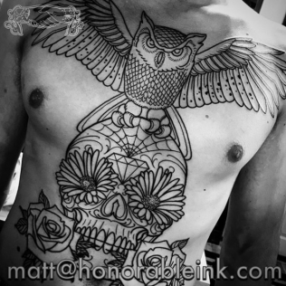 Black-and-white photo of a tattooed bare chest with an owl, flowers, skull, and rose tattoo design
