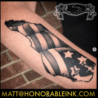 Black and gray Maryland flag tattoo on a forearm, with a star and state silhouette.