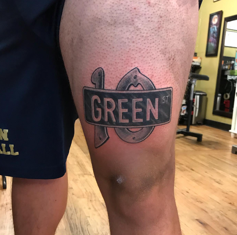 Close-up of a leg with a green tattoo reading “GREEN” in a gym-like indoor setting