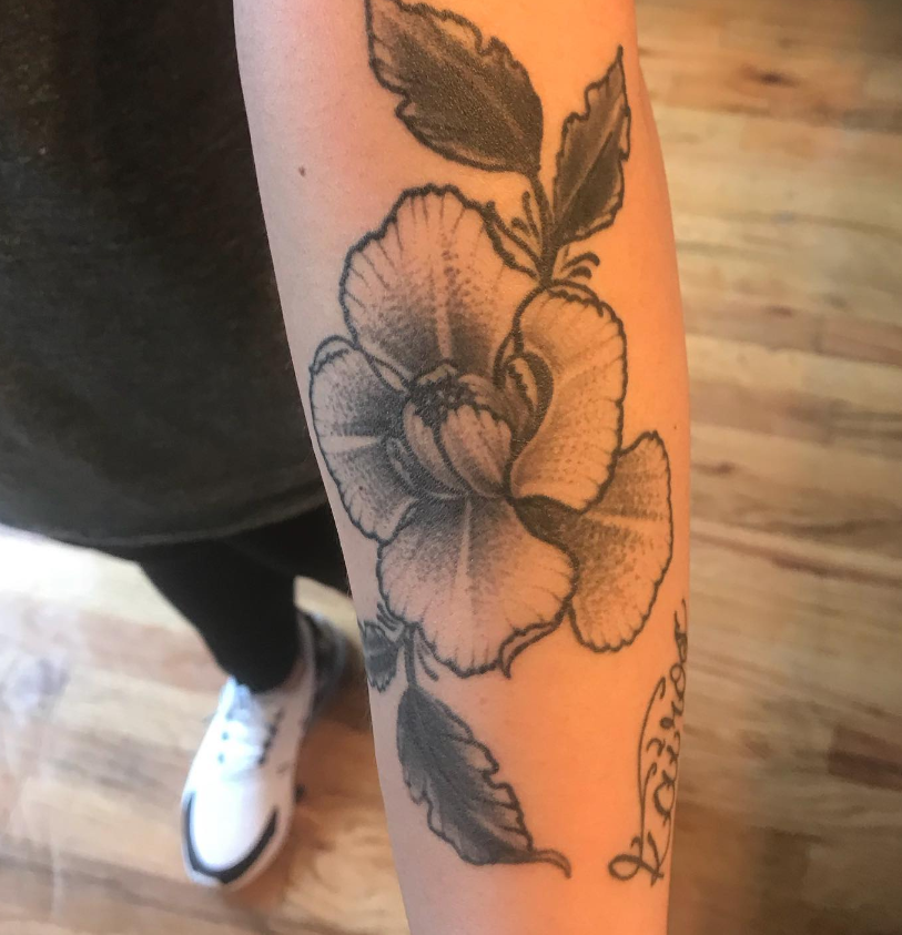 Close-up of a forearm with a black floral tattoo, shown against a wooden floor and sneaker-clad leg.