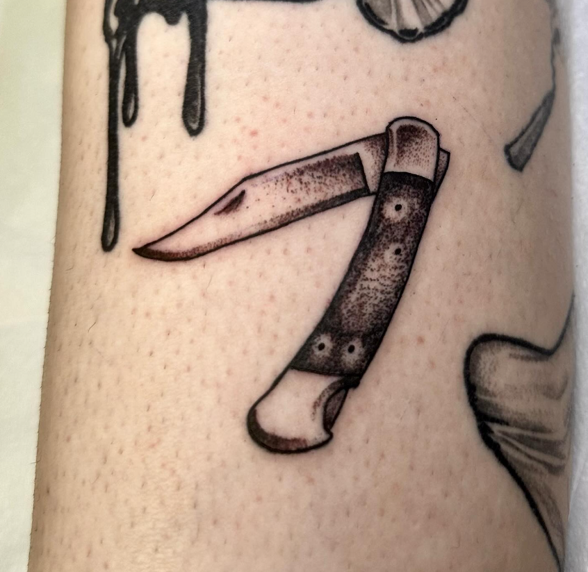 Close-up of a small black-and-gray tattoo of a folding knife on skin