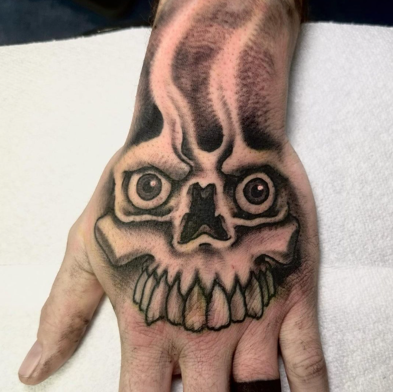 Hand tattoo of a spooky skull face with bulging eyes and sharp teeth in black and pink ink