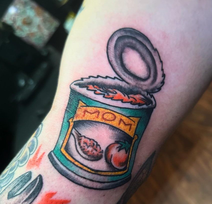 Tattoo of an open can labeled “MOM” with food spilling out, on skin.
