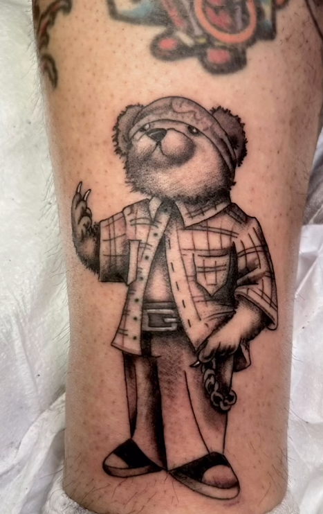Black-and-white tattoo of a cartoon bear in a plaid shirt holding a small object.