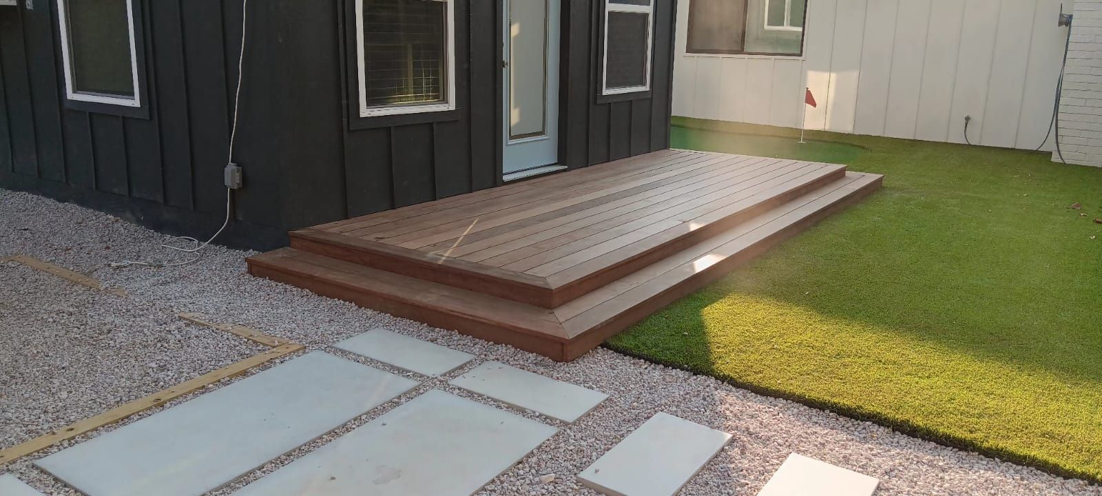 A small brown wooden deck with two steps attached to the side of a modern black building next to a patch of green grass.