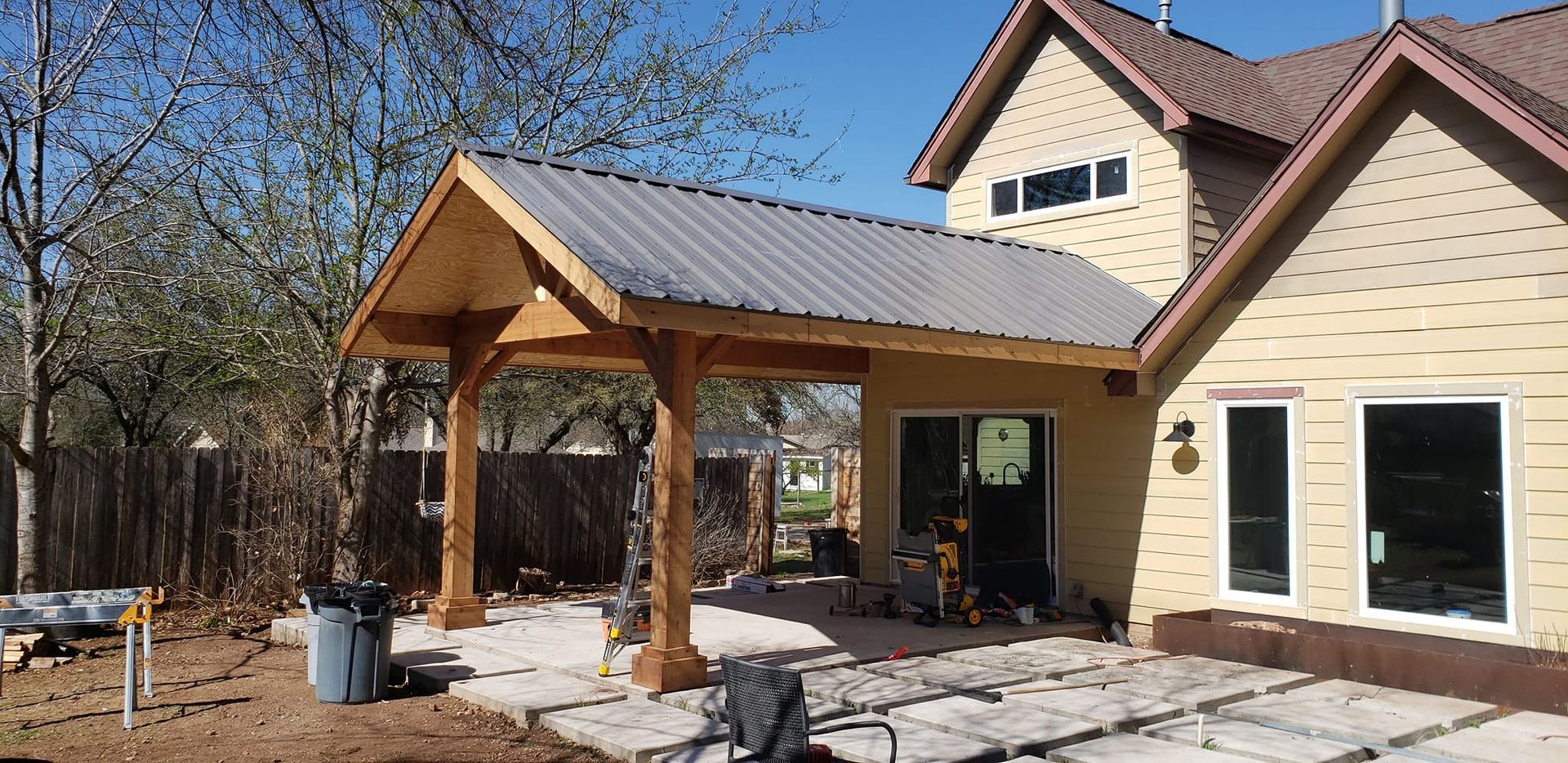 A beige house under construction features a newly built wooden covered patio with a metal roof and a stone-tiled yard.