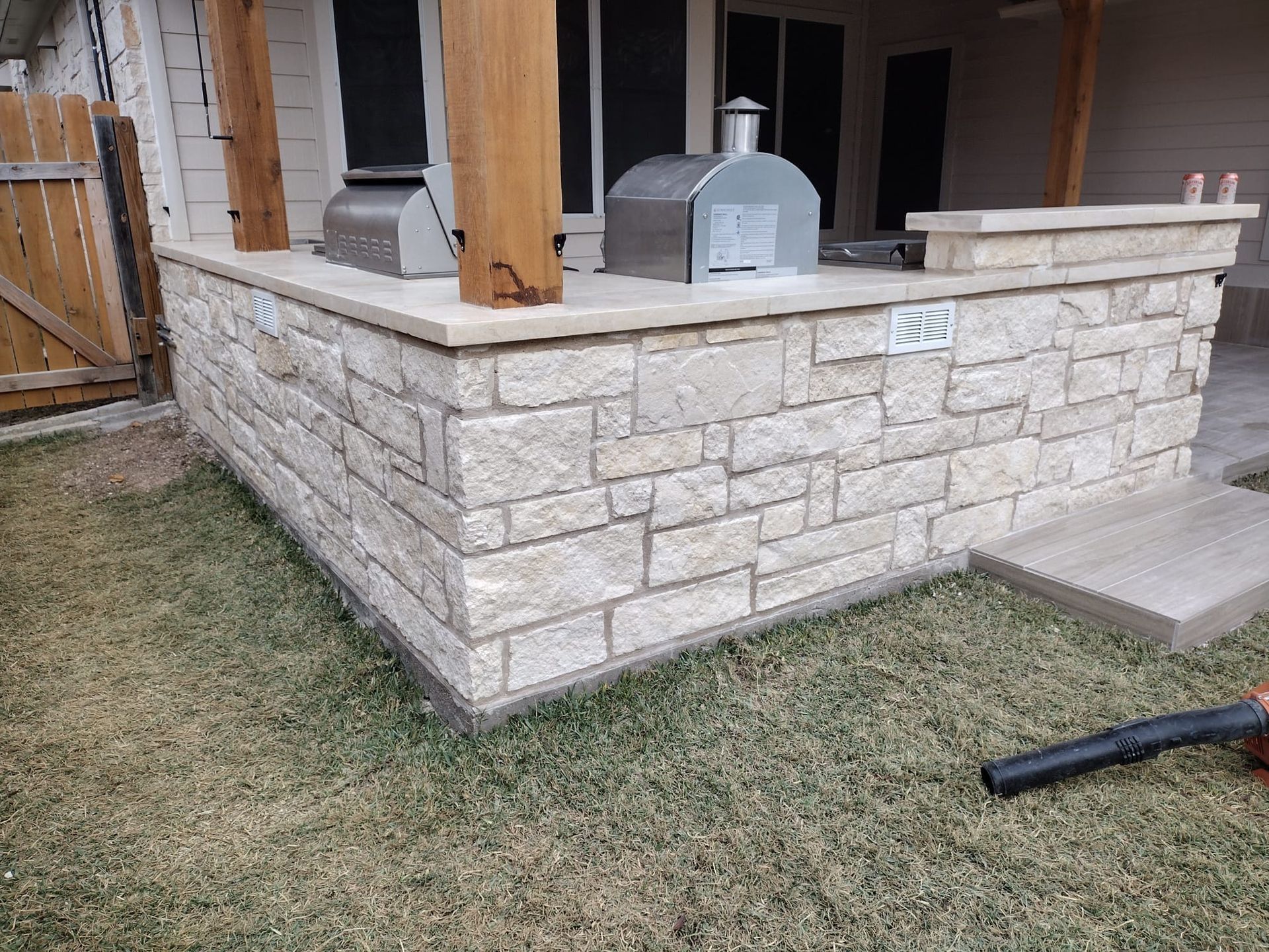 Outdoor kitchen with a stone base, wooden post, and a stainless steel pizza oven on a light-colored countertop.