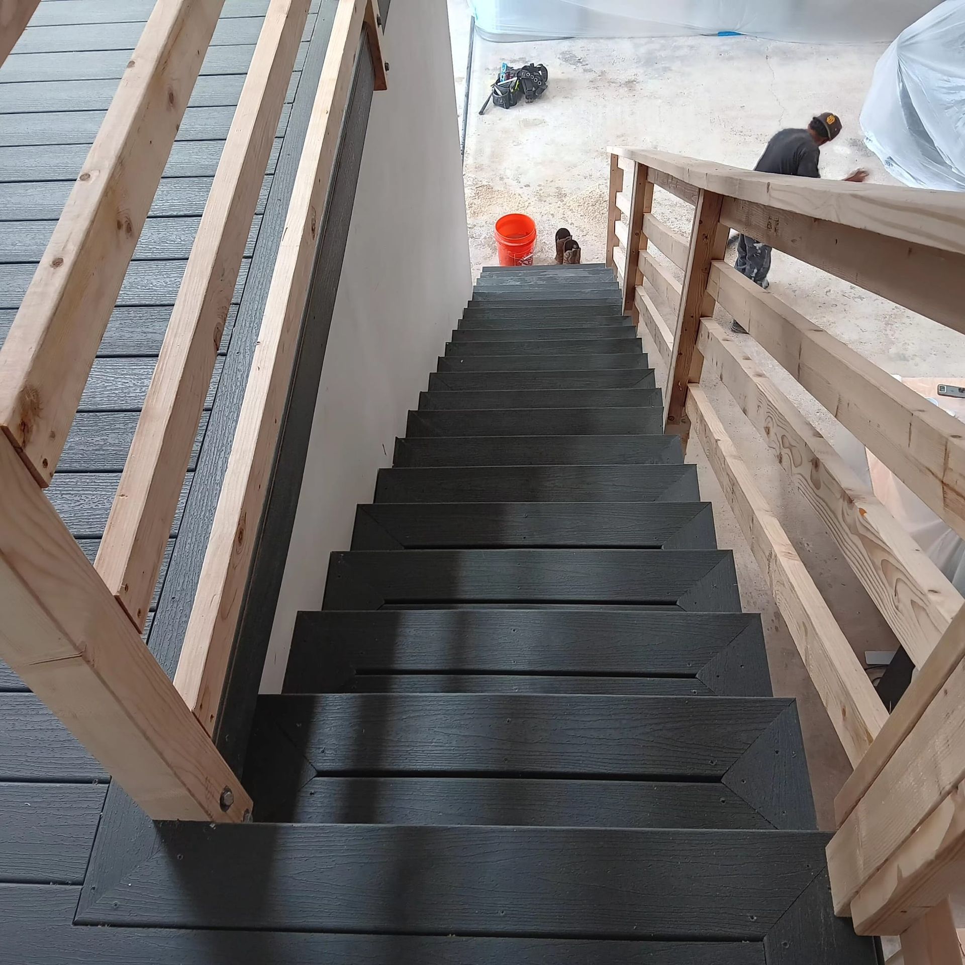 A wooden staircase with dark steps and railings leads down to a construction site with a worker and an orange bucket.