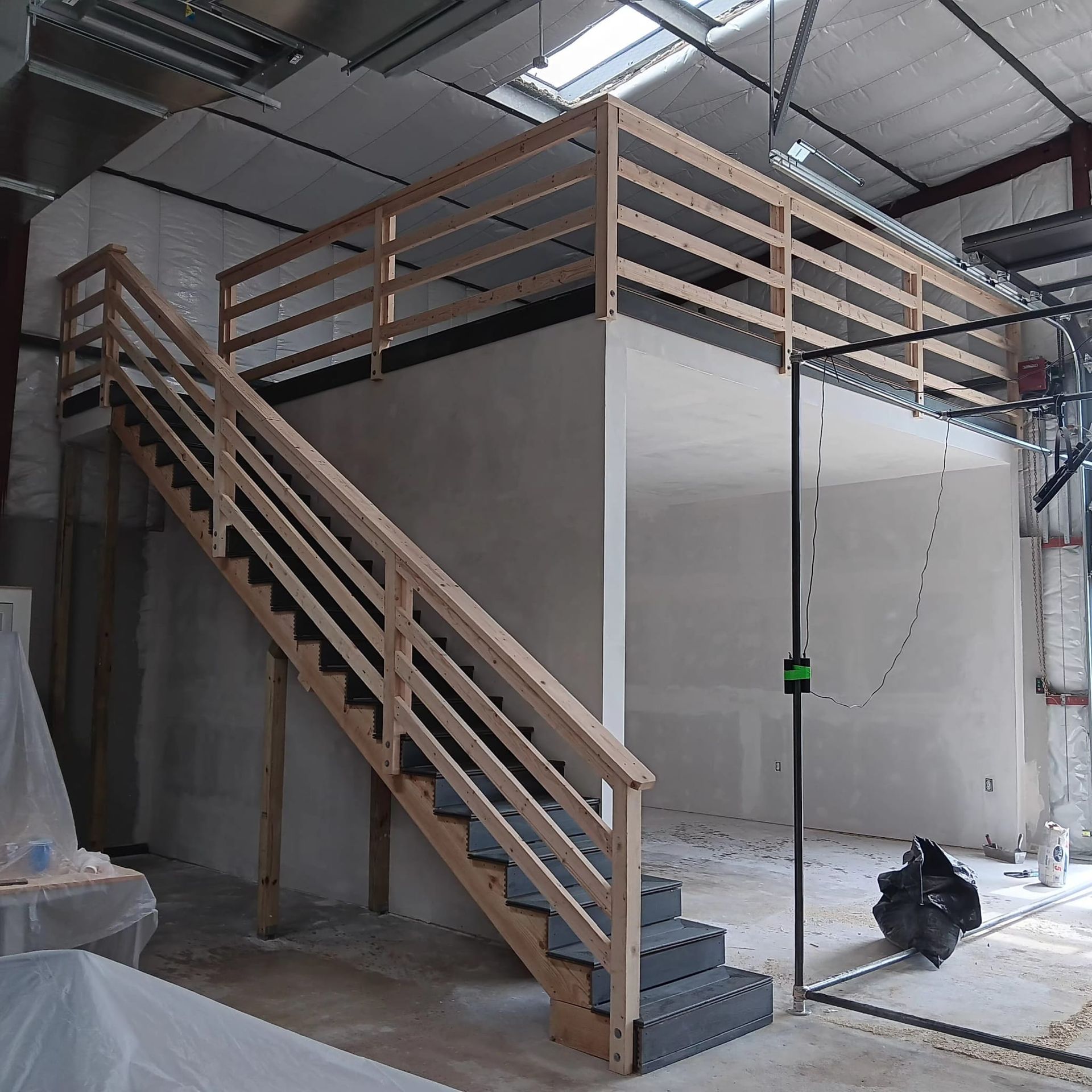 A wooden staircase leads to a loft platform with a railing, located inside a workshop under construction.