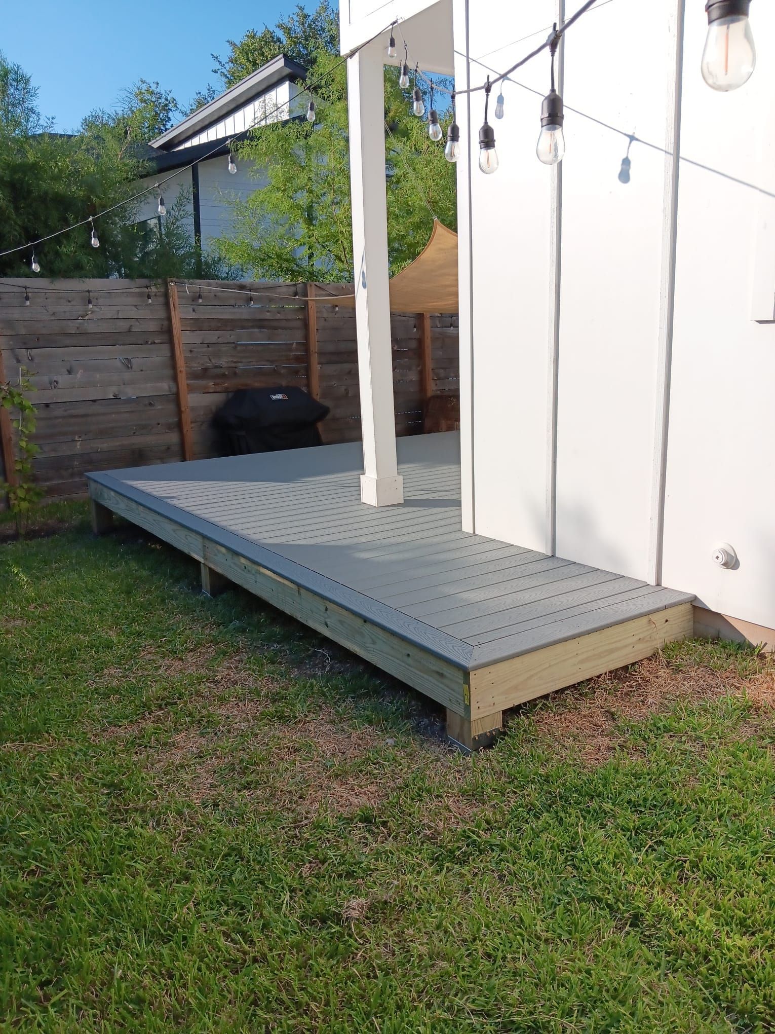 A grey deck with a white support post in a grassy backyard next to a white wall with string lights.