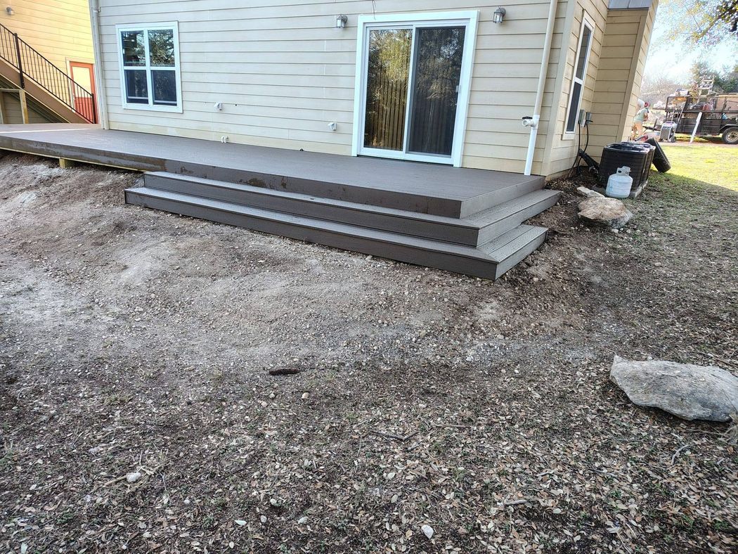 A gray deck with three wide steps leads to a beige house with a sliding glass door and a window, set in a dirt yard. A gray deck with three wide steps leads to a beige house with a sliding glass door and a window, set in a dirt yard.