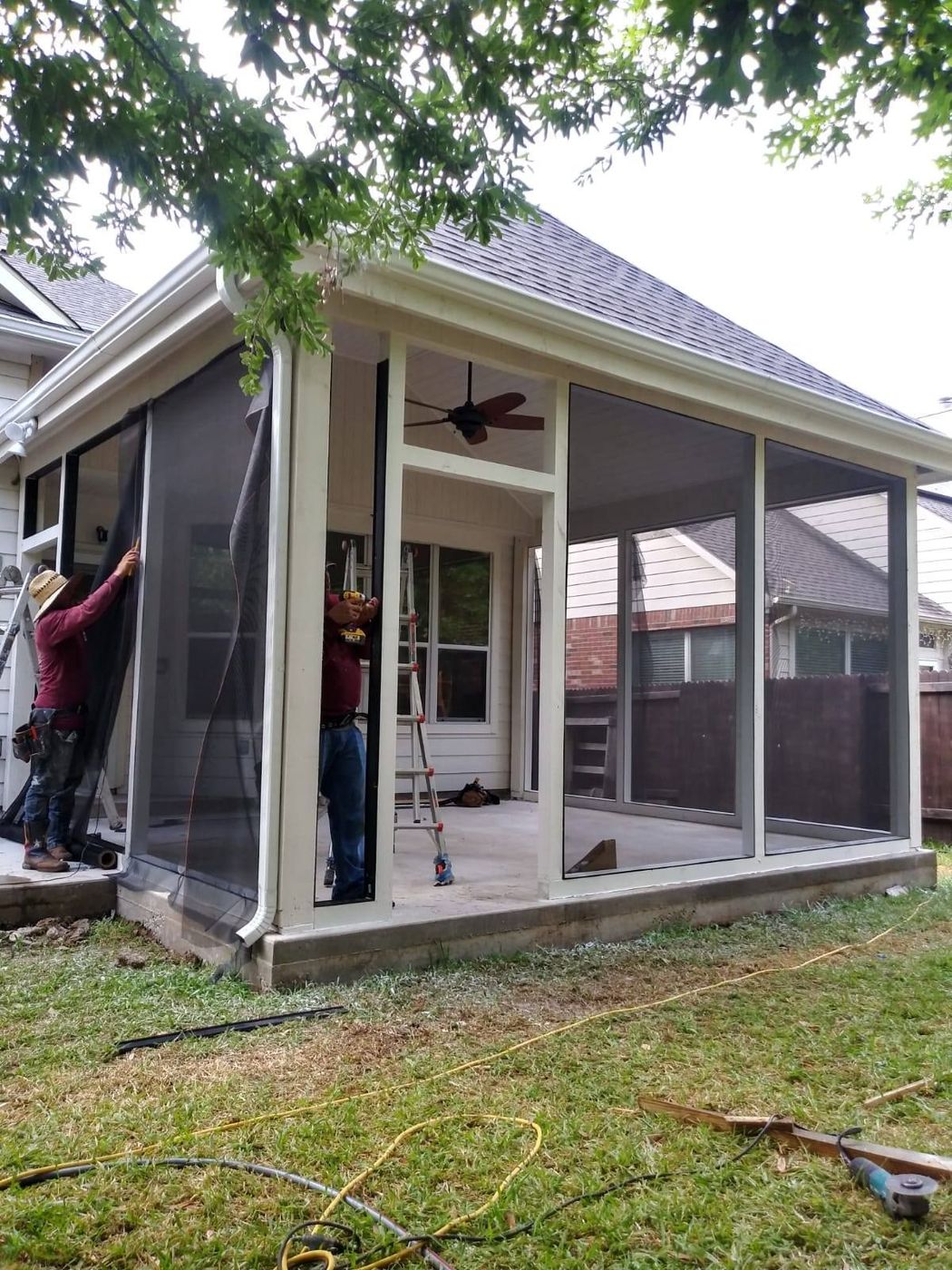 Two workers install large window screens into a cream-colored outdoor gazebo frame on a grassy lawn. Two workers install large window screens into a cream-colored outdoor gazebo frame on a grassy lawn.